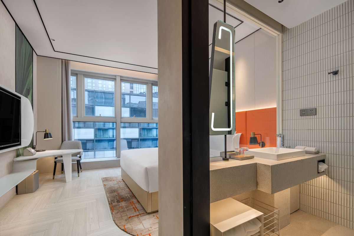 A modern hotel room with a bed, desk, and large window.