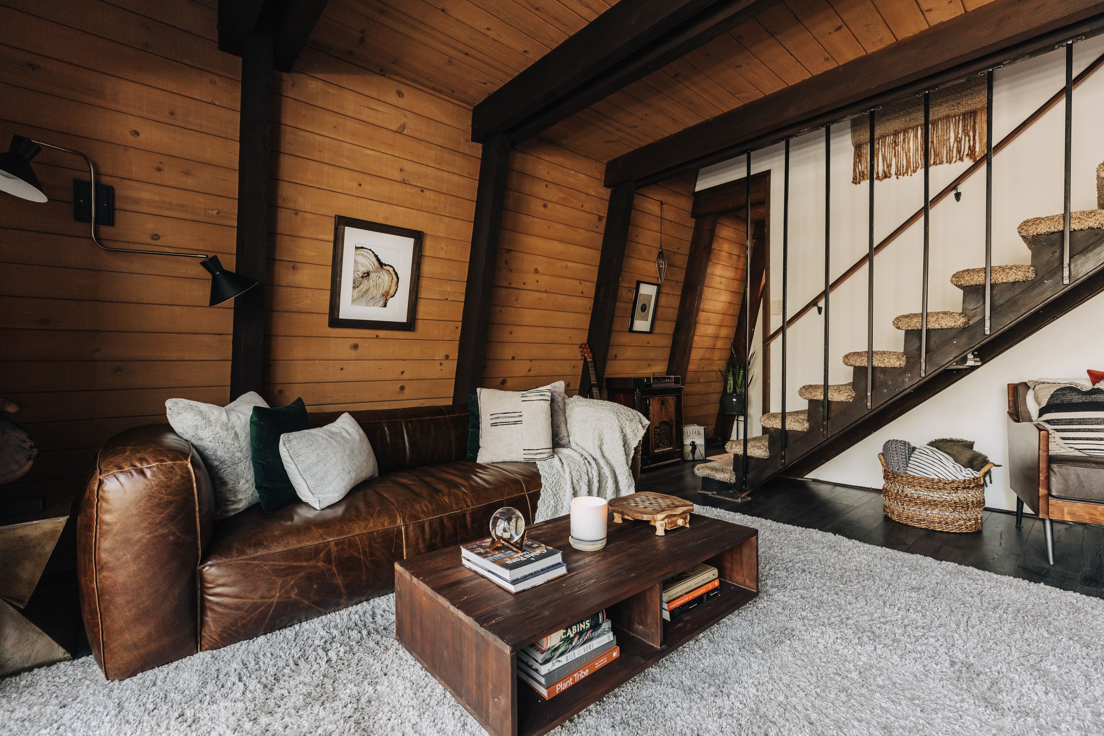 A cozy living room with a brown leather sofa, a wooden coffee table with books, a white rug, and a staircase with wooden steps.