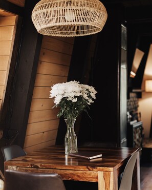 Una mesa de comedor de madera con un jarrón de cristal transparente con flores blancas, una lámpara colgante de mimbre y una libreta.
