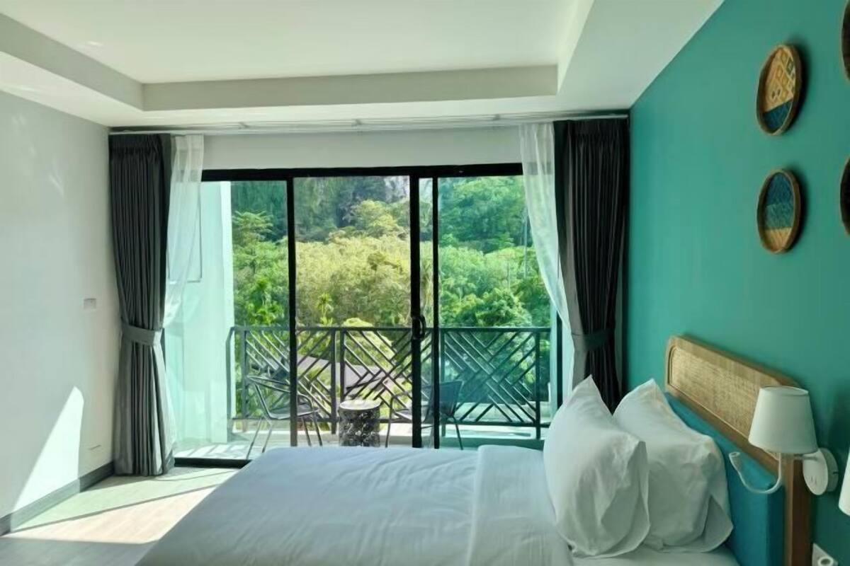 A bedroom with a large bed, a view of greenery through a sliding door, and a balcony with chairs.