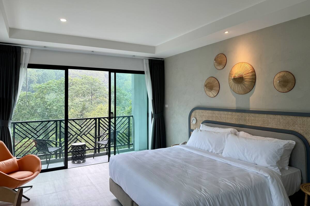 A modern hotel room with a large bed, a balcony with a view of greenery, and decorative wall art.