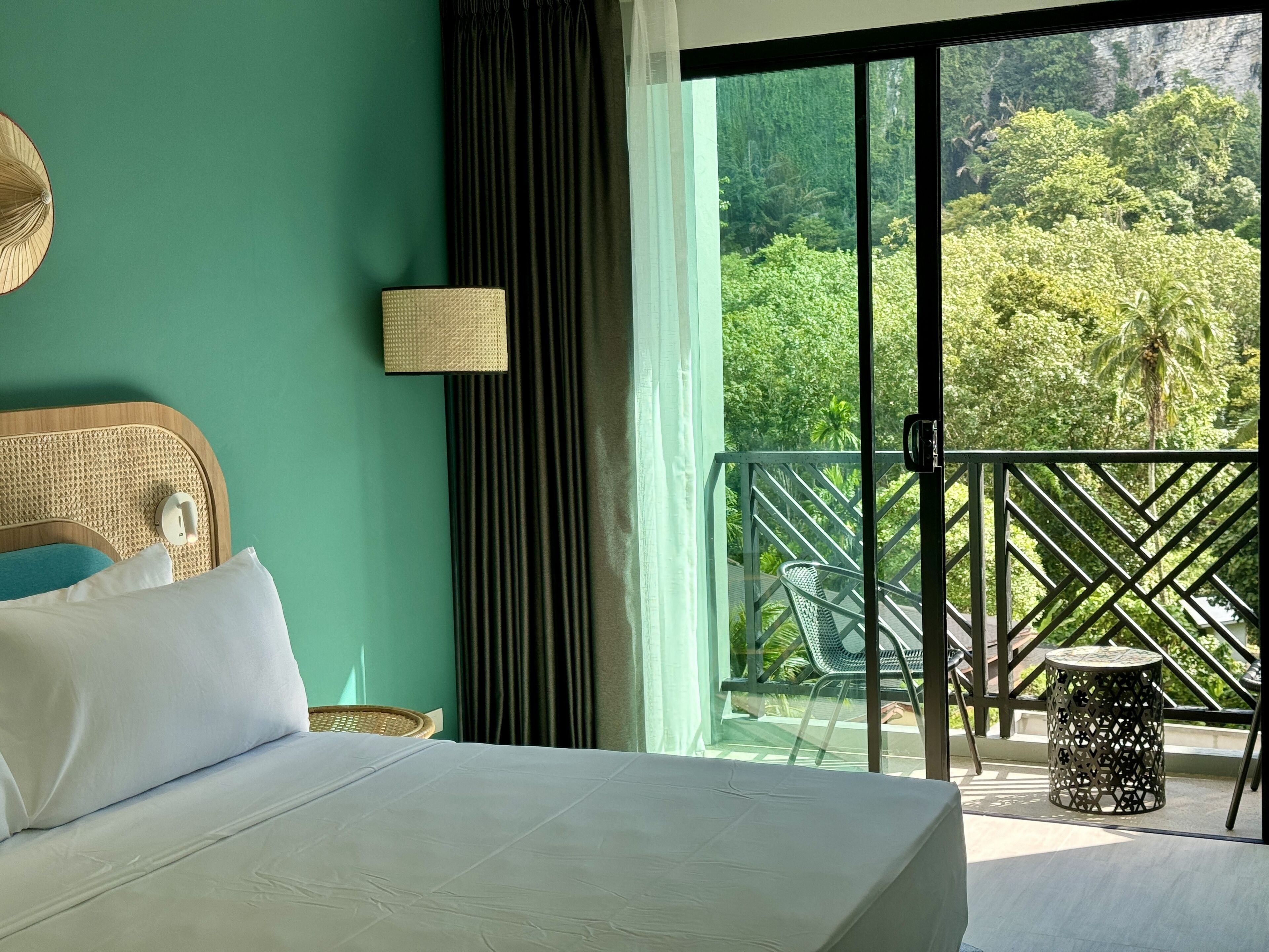 Une chambre d’hôtel avec un lit, une lampe fixée au mur et une vue sur une végétation luxuriante, visible à travers la porte d’un balcon.