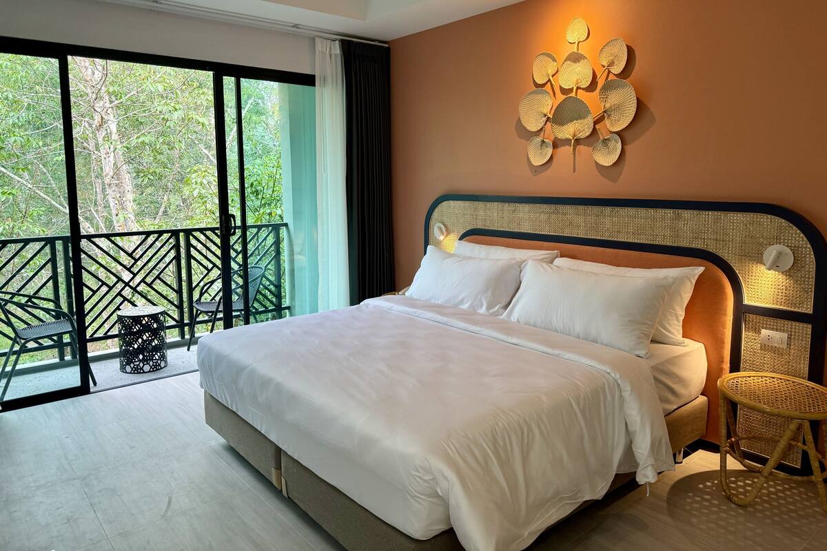 A bedroom with a large bed, a balcony with chairs, and a view of greenery.