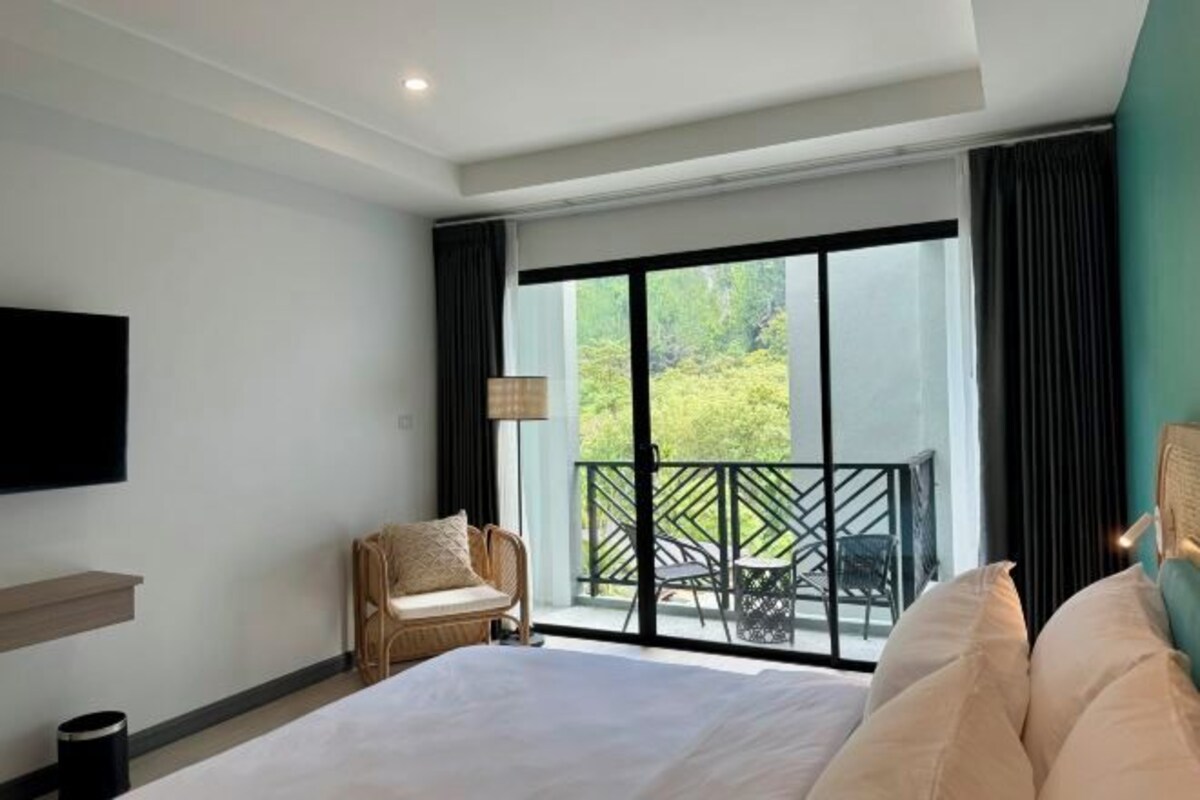 A hotel room with a large bed, a chair, a TV, and a balcony with outdoor furniture.