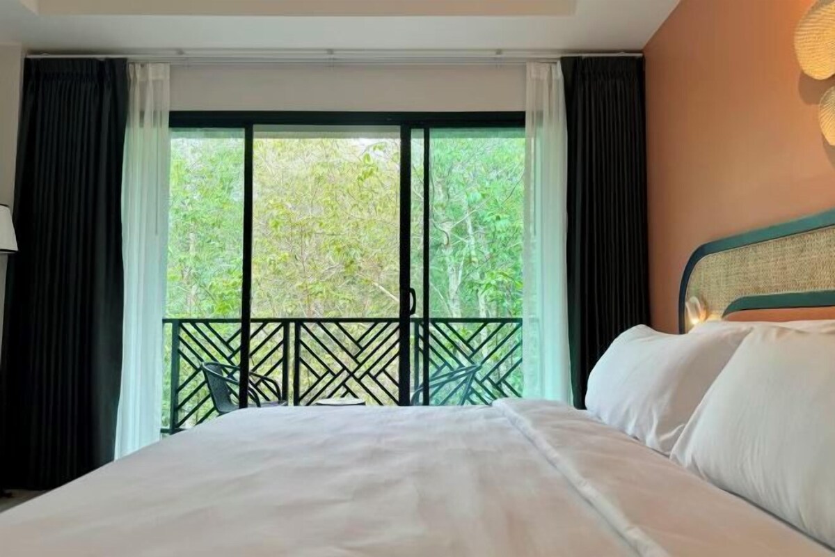 A hotel room with a bed, a balcony with a view of trees, and a window with curtains.