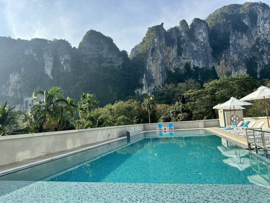 A swimming pool with clear blue water, surrounded by lounge chairs and umbrellas, with a mountainous backdrop.