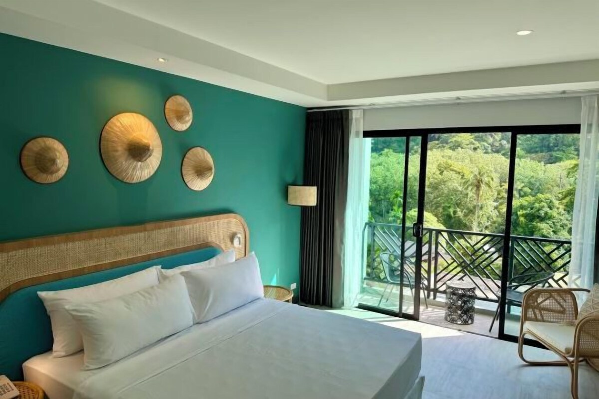 A hotel room with a bed, a view of greenery through large windows, and a balcony with chairs.