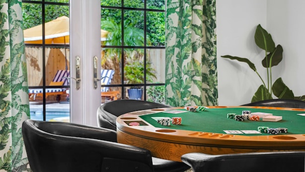Et kasino med en spillebord, pokerchips og spillekort, omgivet af sorte læderstole, med udsigt til grøn vegetation udenfor et vindue med et gittermønster.