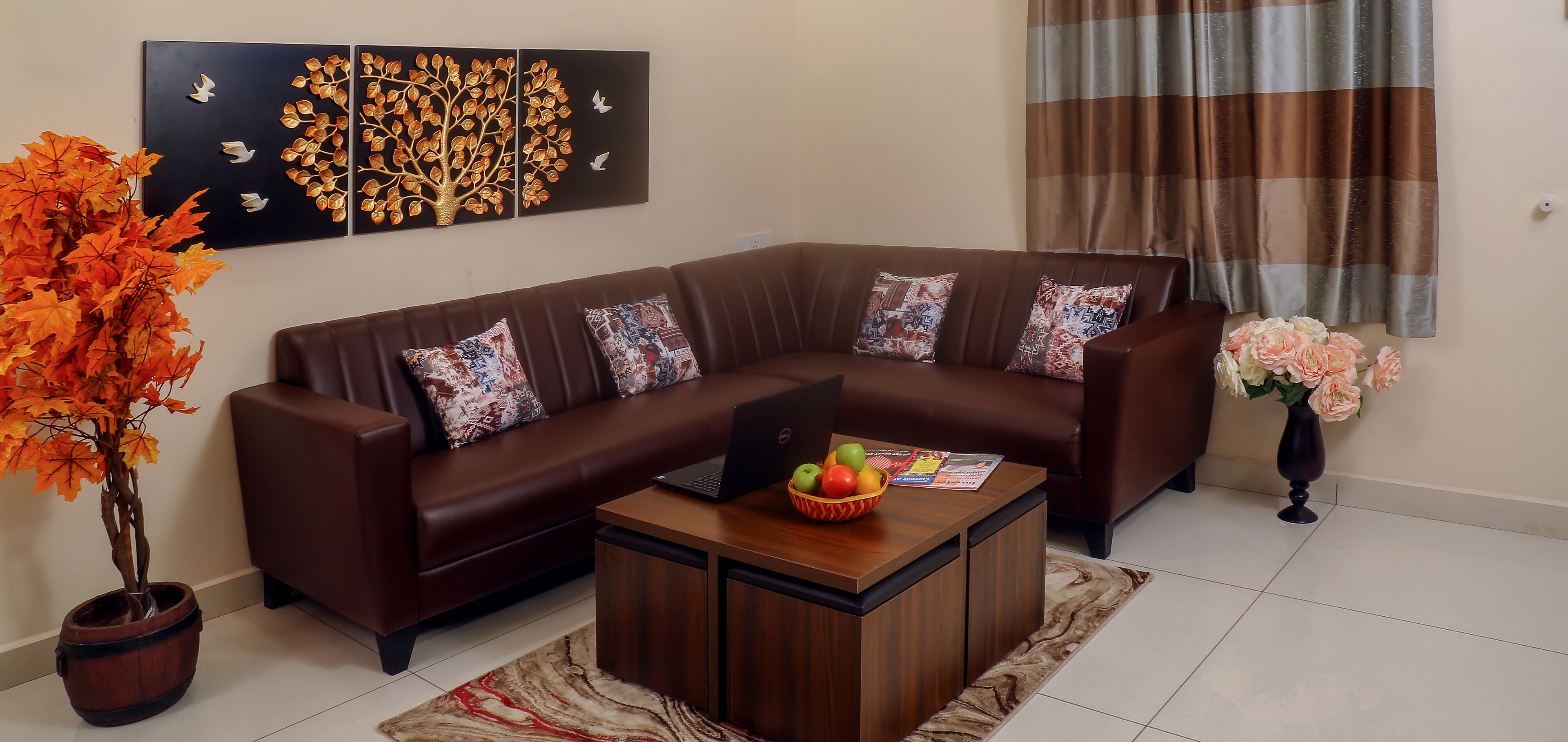 A modern living room with a brown leather sectional sofa, a wooden coffee table with a laptop and fruit bowl, a potted plant, and a wall art piece.