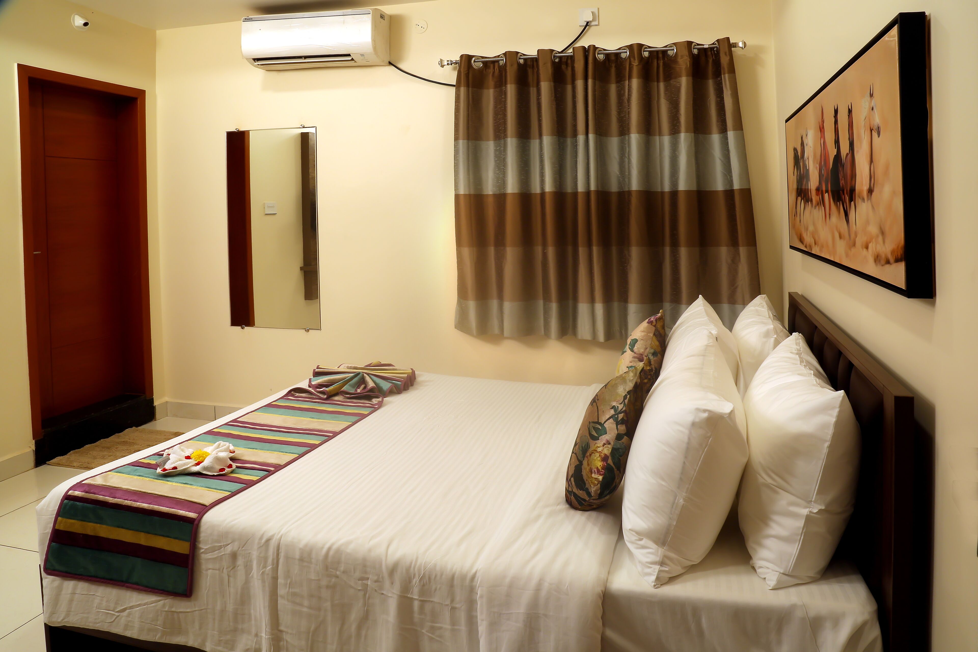A hotel room with a bed, a striped blanket, a pillow, a wall-mounted air conditioner, a curtain, and a painting.