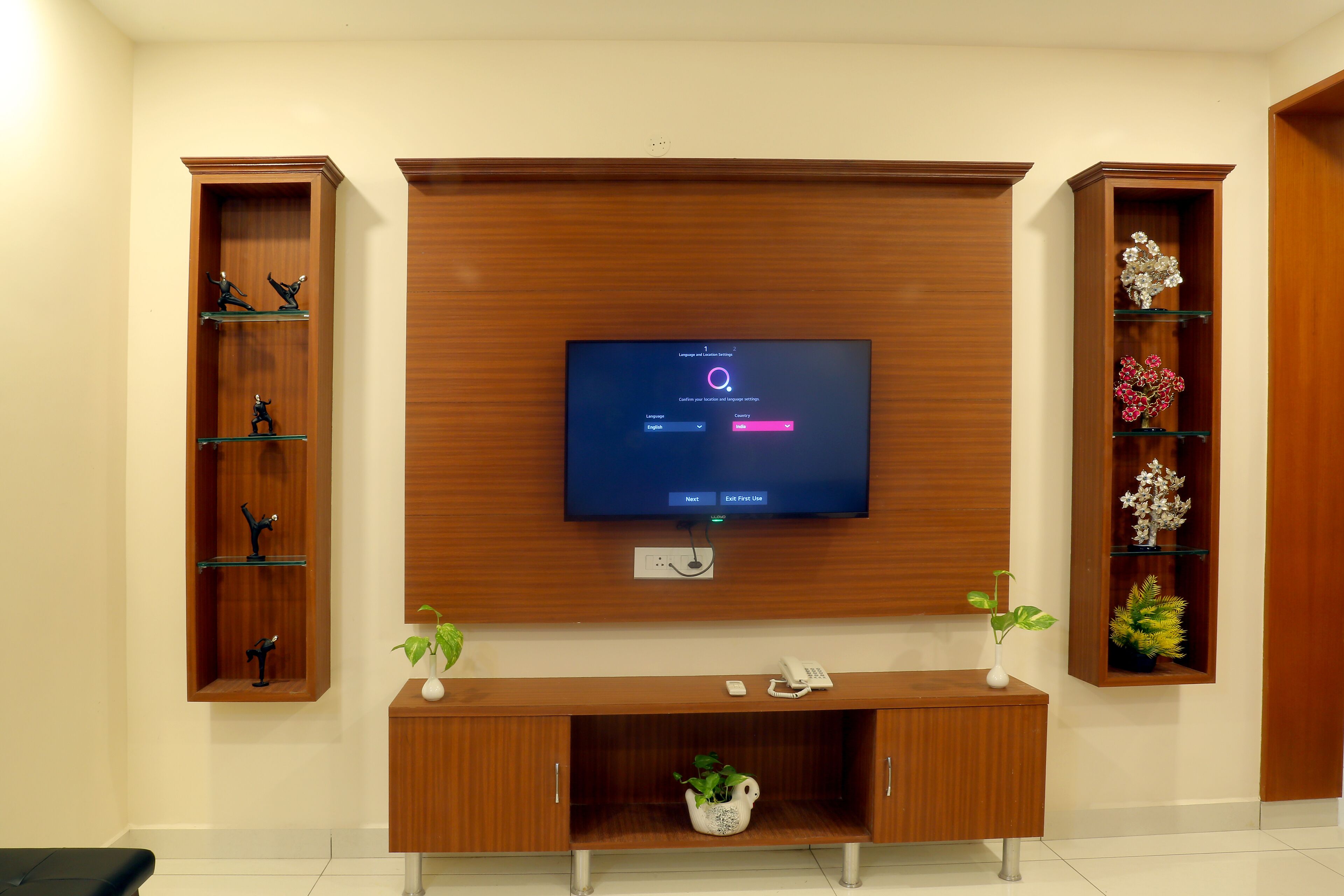 Un salon moderne avec un téléviseur à écran plat fixé à un mur en bois, une console en bois ornée d’un vase et d’une plante, et deux étagères en bois sur lesquelles sont disposés des objets décoratifs.