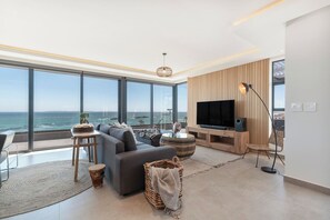 A modern living room with a large TV, comfortable seating, and a view of the ocean.