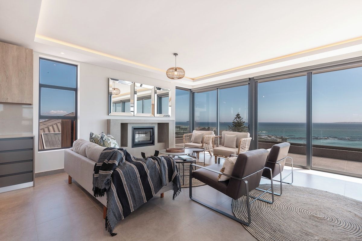 A modern living room with a sofa, armchairs, a coffee table, and a fireplace. Large windows offer a view of the ocean.