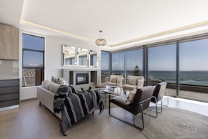A modern living room with a sofa, armchairs, a coffee table, and a fireplace. Large windows offer a view of the ocean.