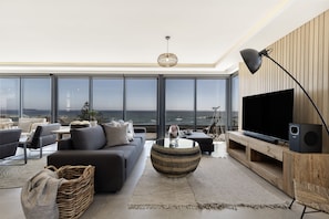 A modern living room with a large TV, comfortable seating, and a view of the ocean.