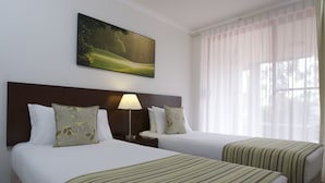 A hotel room with two beds, a nightstand, a lamp, a painting, and a window with sheer curtains.