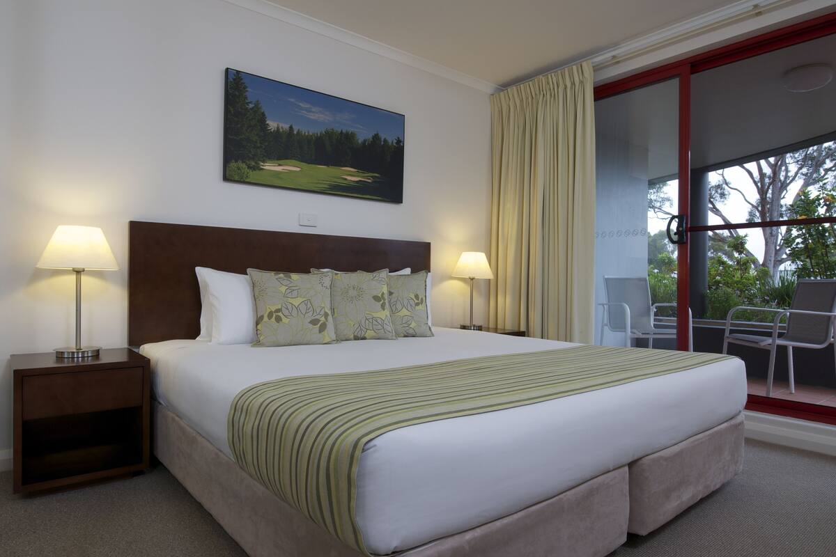A hotel room with a large bed, two bedside lamps, a painting of a golf course, and a view of trees through a sliding glass door.