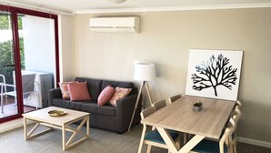 A living room with a sofa, a coffee table, a dining table with chairs, a floor lamp, and a wall-mounted air conditioner.