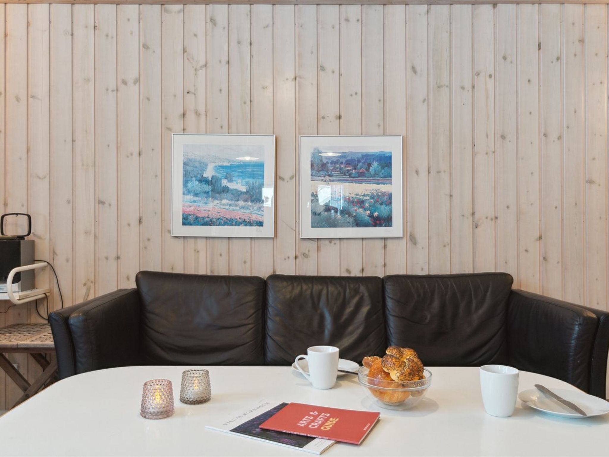 Une pièce avec un mur lambrissé en bois, deux tableaux encadrés, un canapé en cuir noir, une table blanche sur laquelle se trouve un saladier rempli de viennoiseries, un livre rouge et deux bougies allumées.