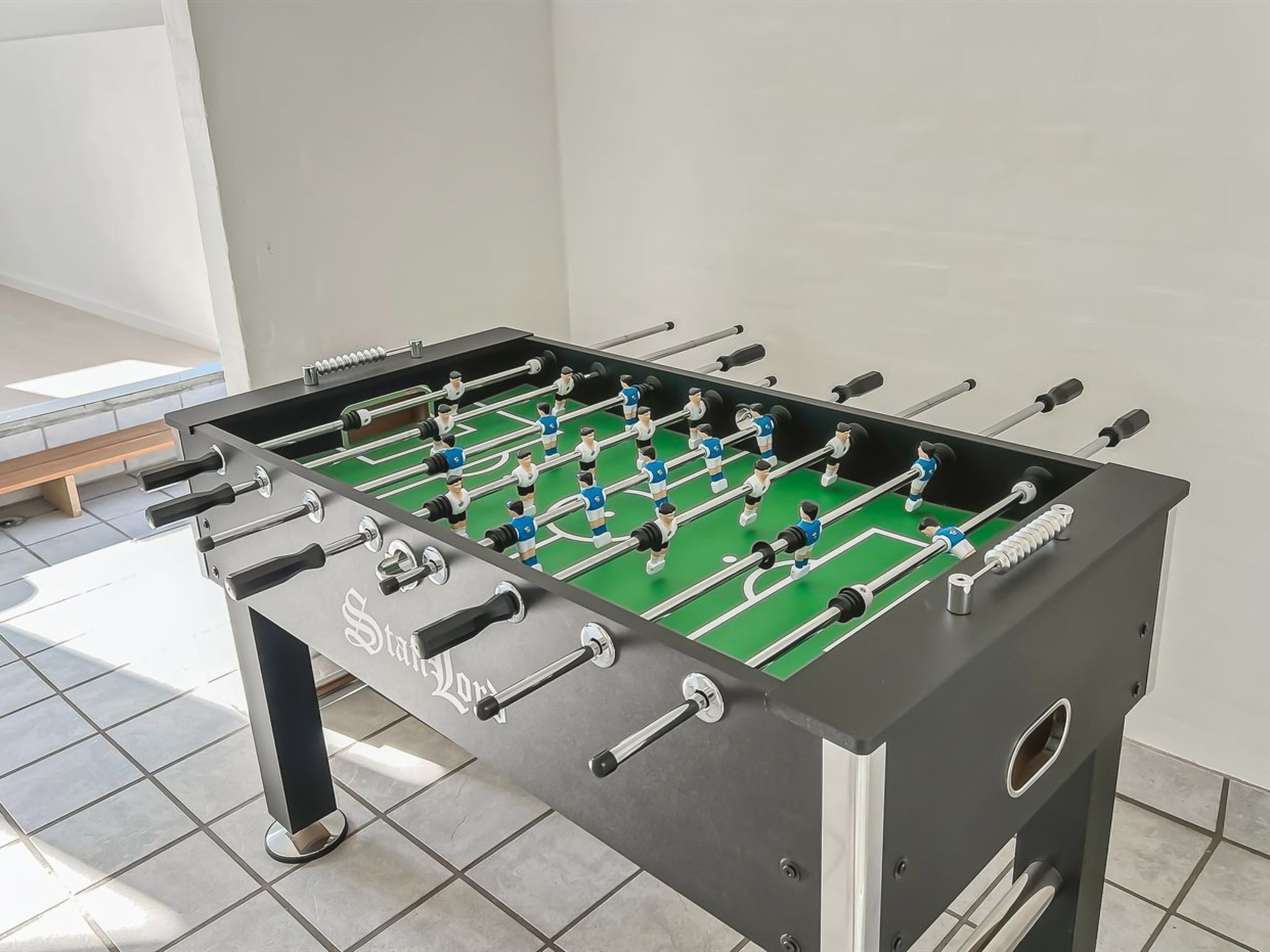 A foosball table with a metallic finish and a green playing surface, set against a tiled floor in a room with a white wall.