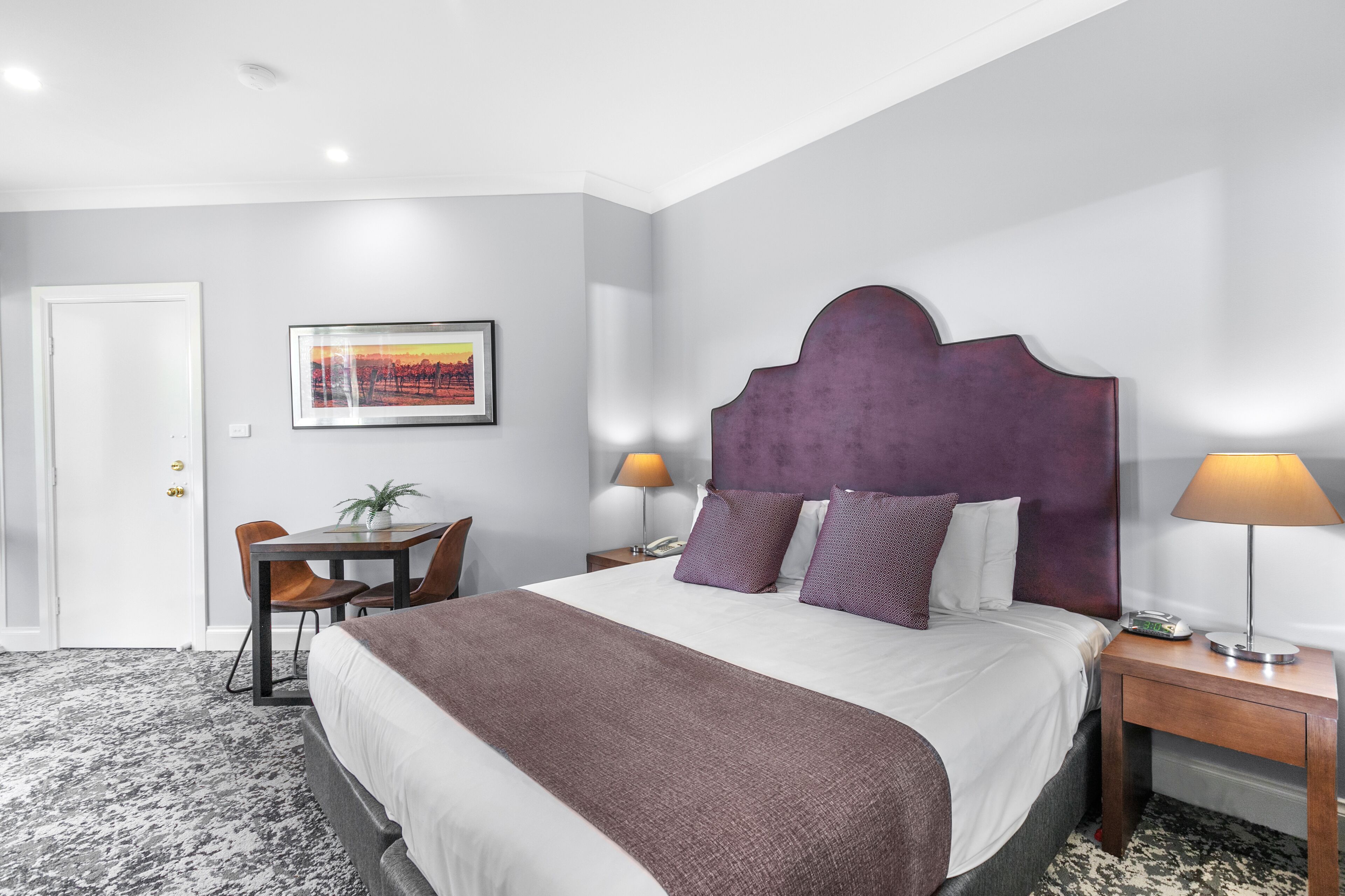 A hotel room with a large bed, a desk with a chair, a small table with a plant, and a framed picture on the wall.