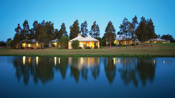 A lakeside hotel with multiple buildings, a calm lake reflecting the structures, and a clear evening sky. - Club Wyndham Pokolbin Hill (Pokolbin)