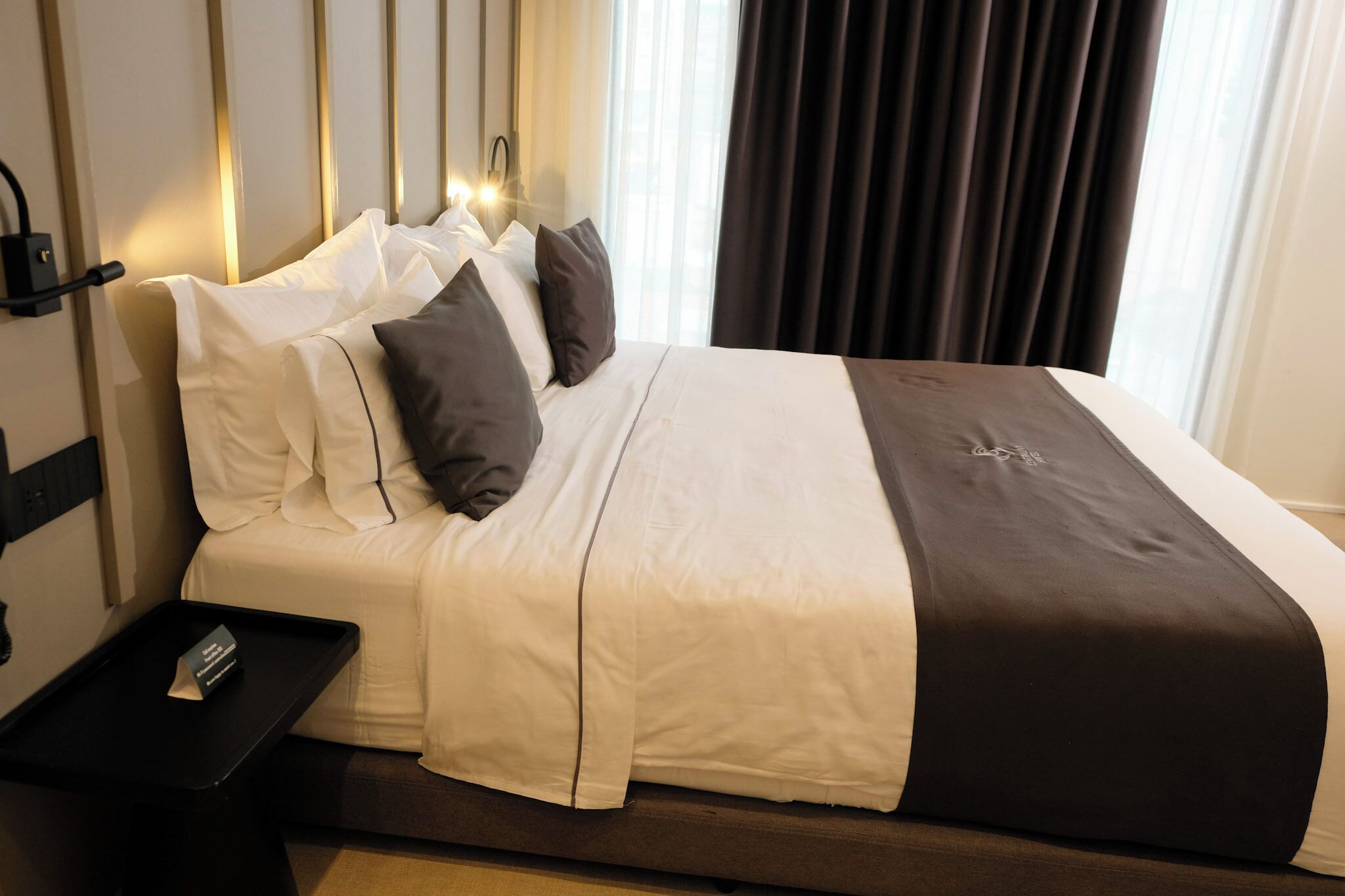 Quarto de hotel com cama bem arrumada, mesa de cabeceira com bandeja e janela com cortinas.