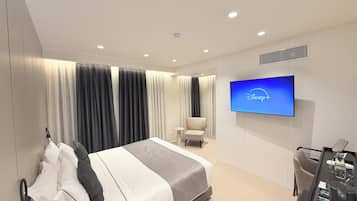 Una habitación de hotel moderna con una cama grande, un televisor de pantalla plana mostrando el logotipo de Disney+, una zona de estar con una silla y una mesita con un hervidor y una taza.