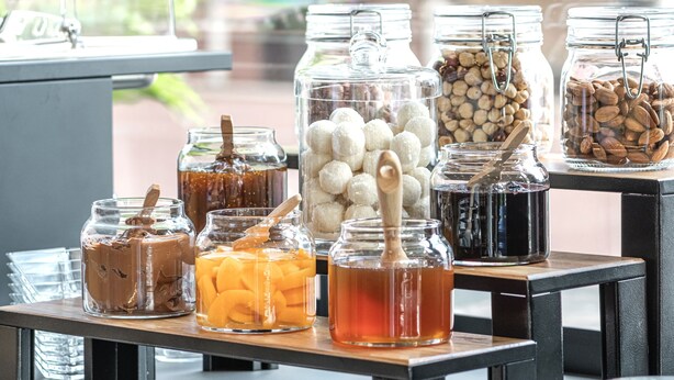 Un bufé con diversos alimentos en frascos y recipientes, como frutos secos, frutas y postres, exhibido sobre una barra con platos y una máquina de café.