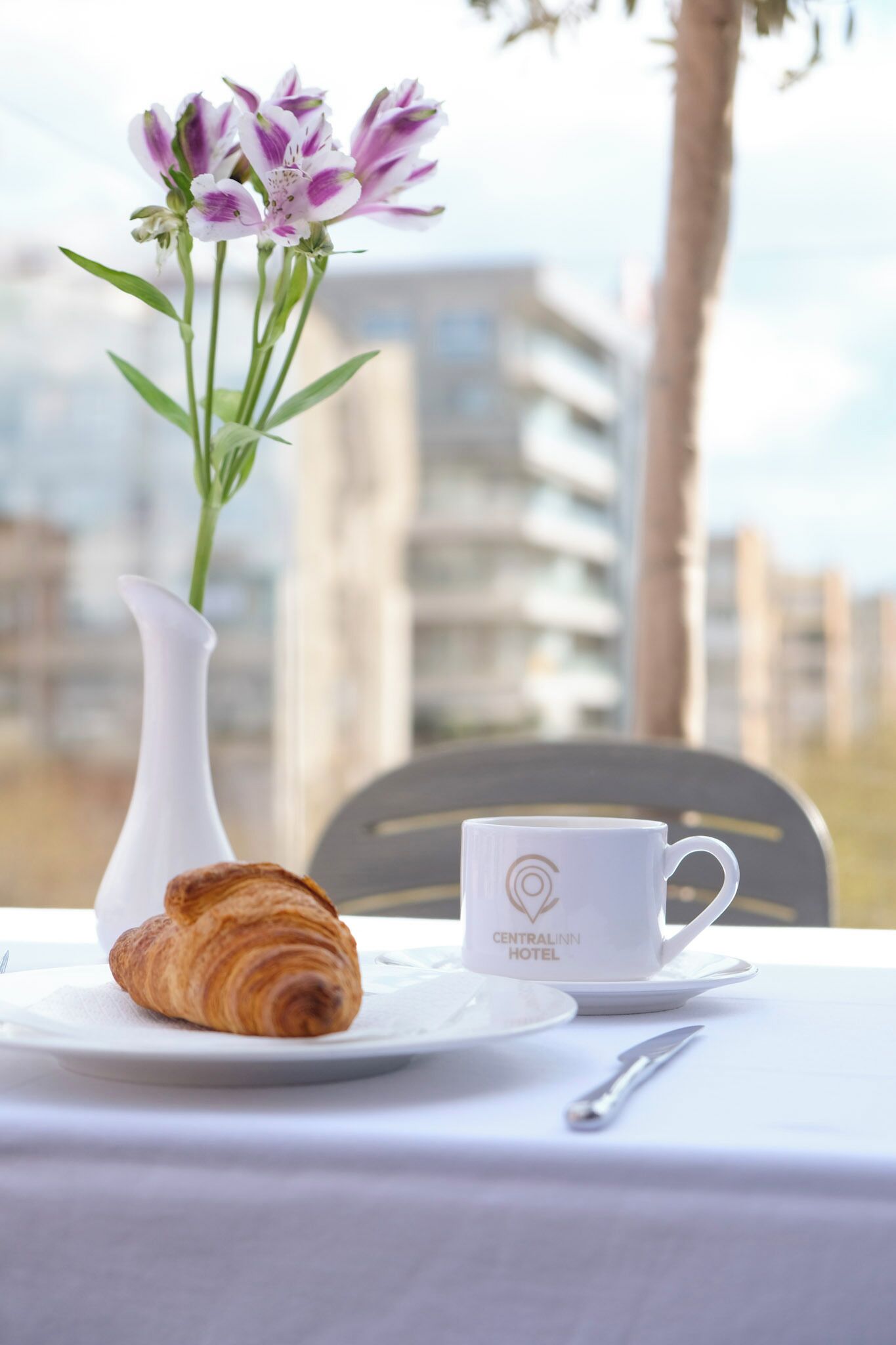 Uma mesa com um croissant, uma chávena branca com o logótipo 'CENTRALINN HOTEL' e um vaso com flores.