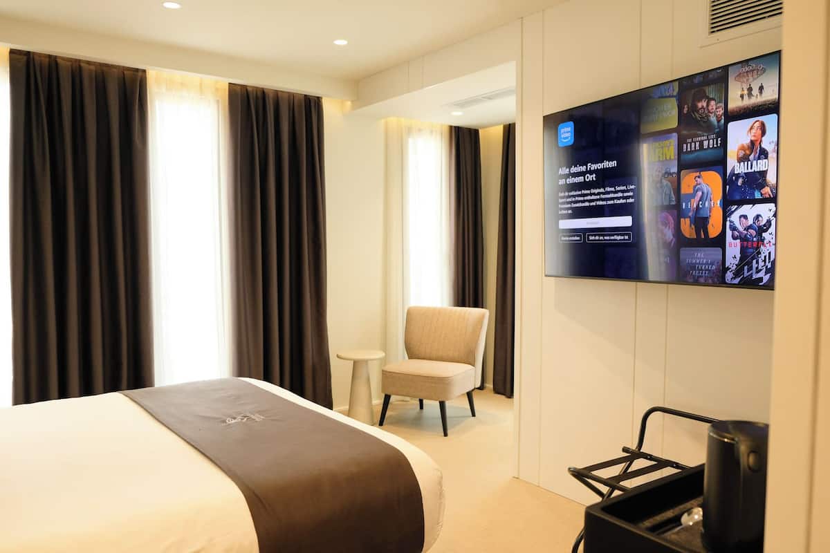 A hotel room with a bed, a chair, a small table, and a wall-mounted TV displaying movie posters.