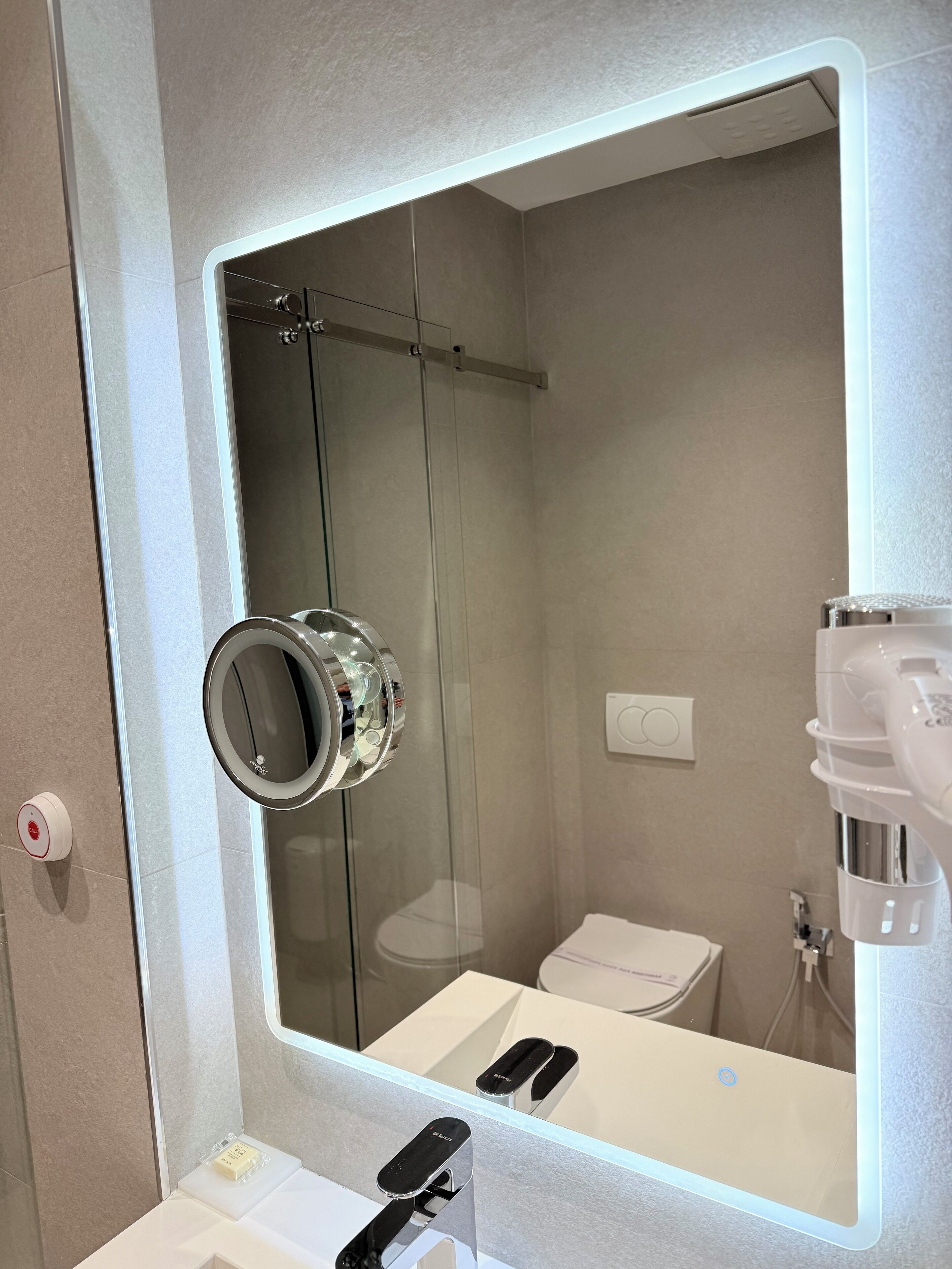 Un baño moderno con un espejo grande, cabina de ducha de vidrio, inodoro y lavamanos con dispensador de jabón.