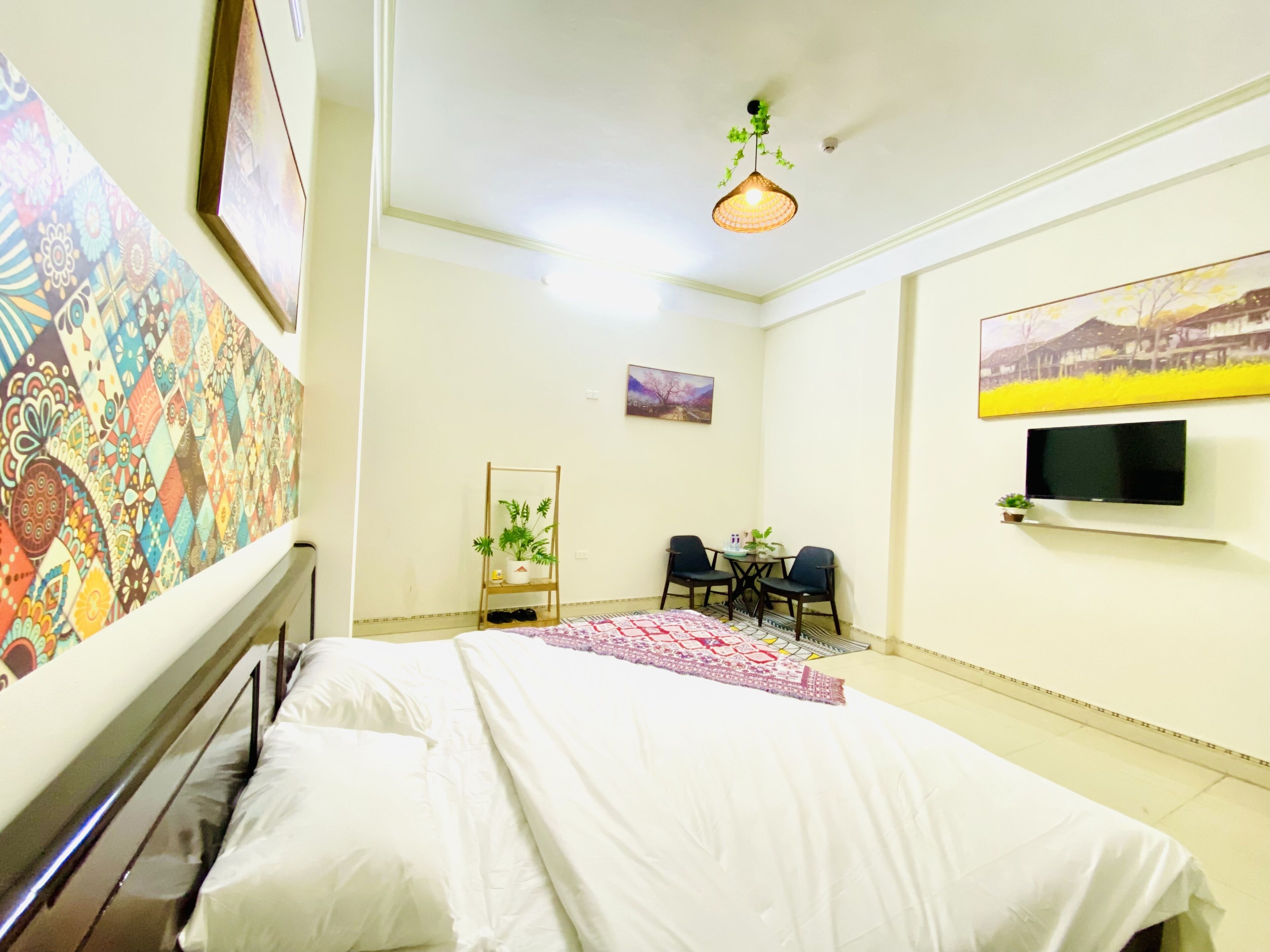 A hotel room with a bed, a small table with chairs, a TV mounted on the wall, and a hanging light fixture.