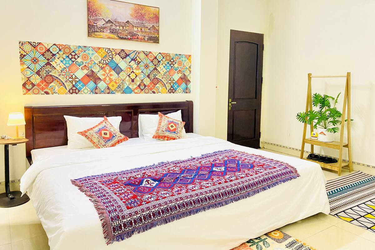 A neatly made bed with patterned pillows and a colorful blanket, a small table with a lamp, a wooden shelf with a plant, and a door in a room with a patterned wall hanging above the bed.
