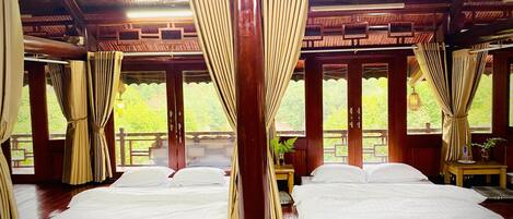 A room with two beds, wooden floors, and large windows with curtains.