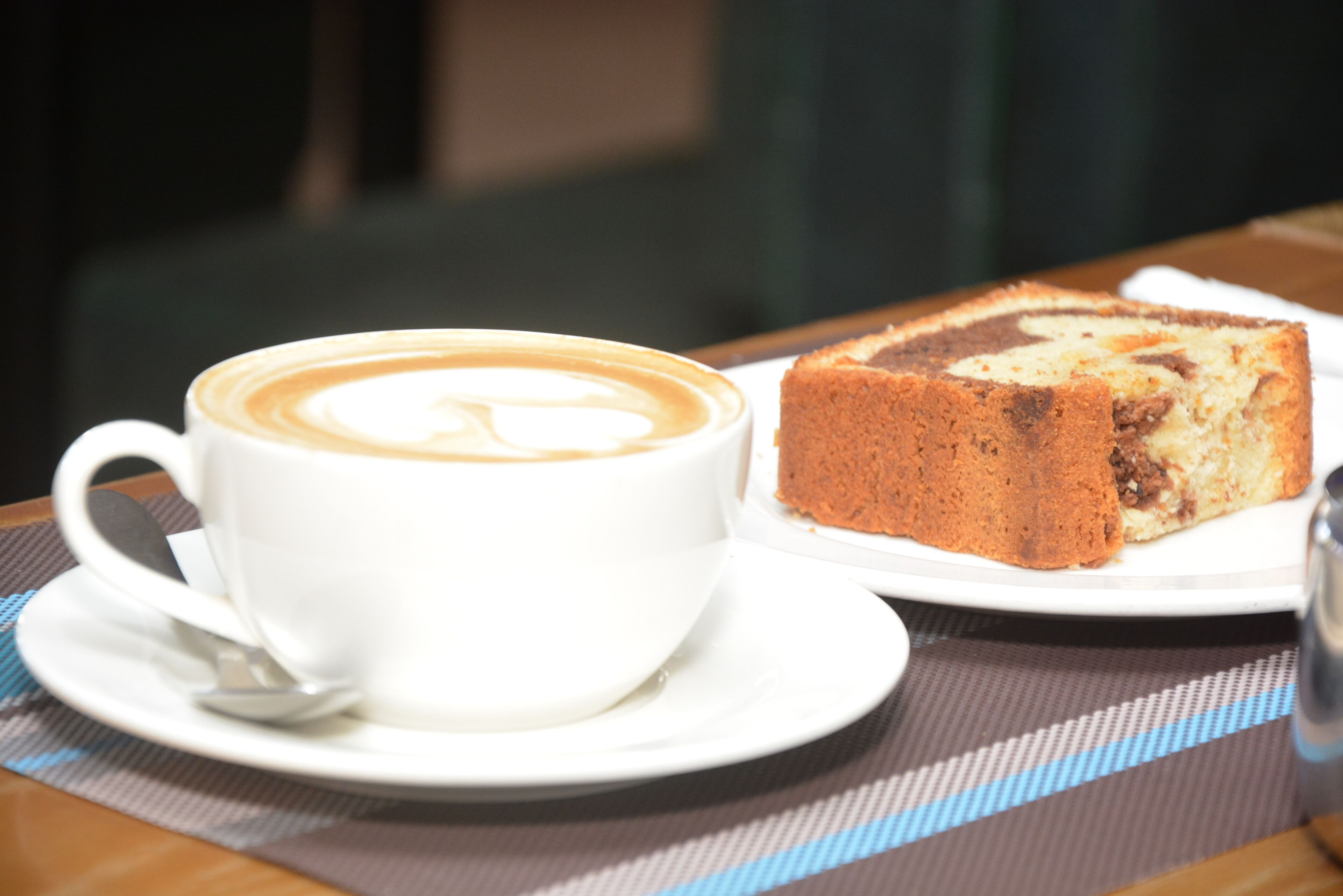 A white cup of coffee with latte art on a white saucer, and a slice of cake on a white plate.