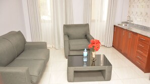Sala de estar con un sofá gris, una mesita con una botella y flores, y una zona de cocina con armarios de madera y un lavamanos.