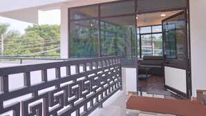 Varanda com guarda-corpo de metal geométrico, porta de vidro para o interior e vista para o verde lá fora.