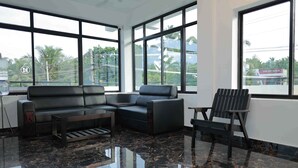 Quarto moderno com sofá de couro preto, uma mesinha e uma cadeira. Janelas amplas oferecem vista para palmeiras e edifícios.