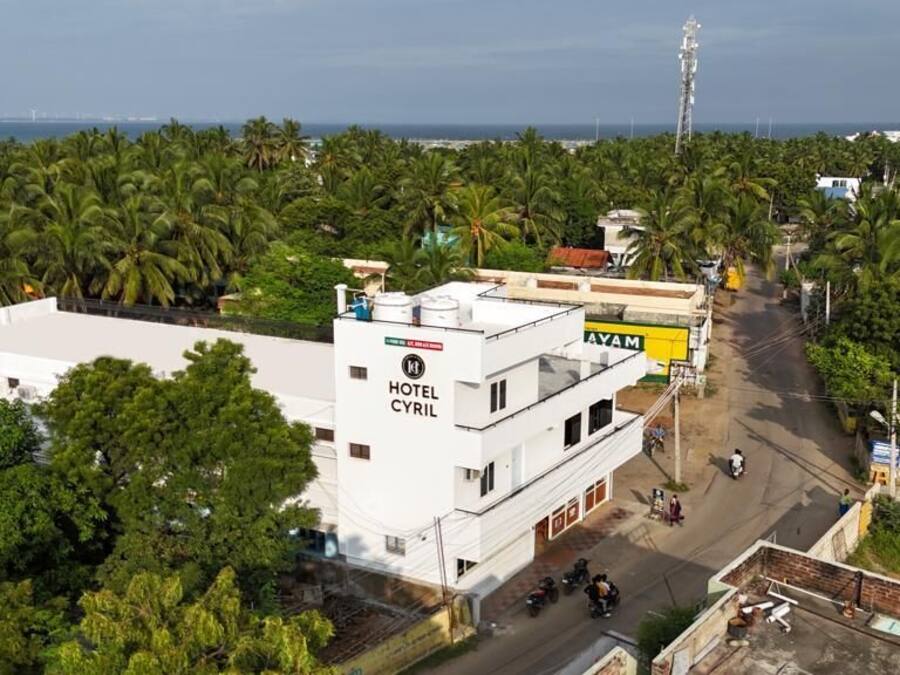 A white hotel named "HOTEL CYRIL" with a signboard, surrounded by palm trees and a road with vehicles.