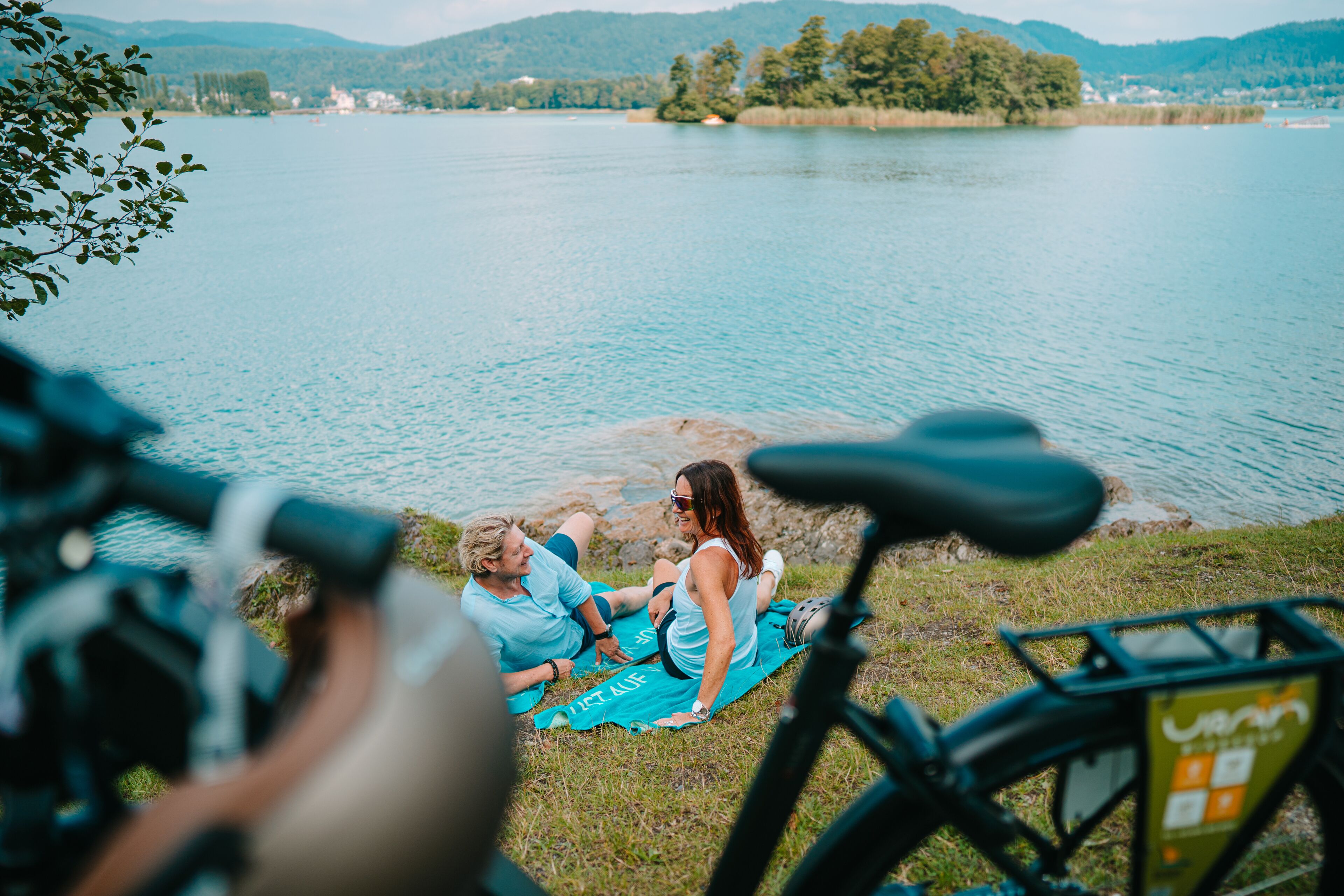 A lake with a bicycle in the foreground and a person sitting on a towel.