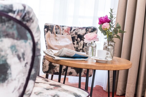 A room with a floral-patterned armchair, a wooden side table with a newspaper, a white mug, and a vase with pink flowers. - WUNDERs Ferienpension  (Poertschach am Woerthersee)