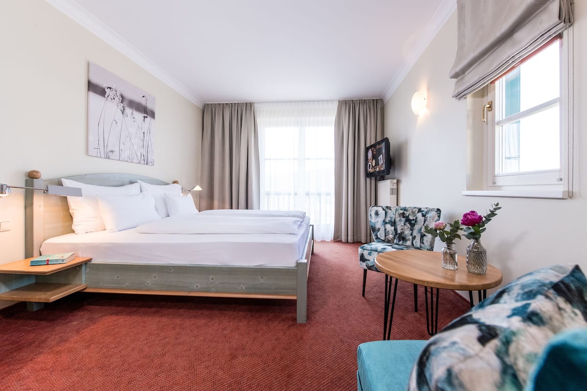 A hotel room with a bed, a chair, a small table with flowers, and a TV on the wall.