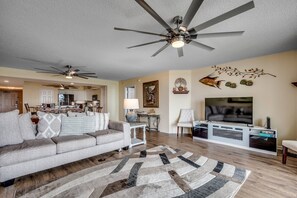 A living room with a sofa, a TV, ceiling fans, a dining area, and a decorative wall piece.