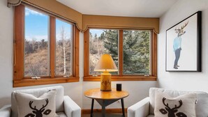 A room with two white armchairs, a small wooden table with a lamp, and a window with a view of trees.