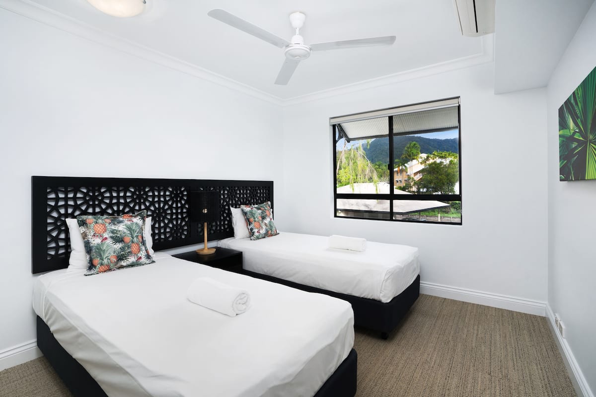 A hotel room with two beds, a ceiling fan, a window with a view of the outdoors, and a decorative wall panel.