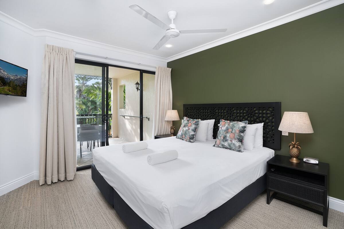 A bedroom with a large bed, two bedside lamps, a ceiling fan, and a view of the outdoors.