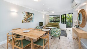 A modern living room with a wooden dining table, green cushioned chairs, a sofa, and a coffee table. There is a painting on the wall, a ceiling fan, and an air conditioning unit.