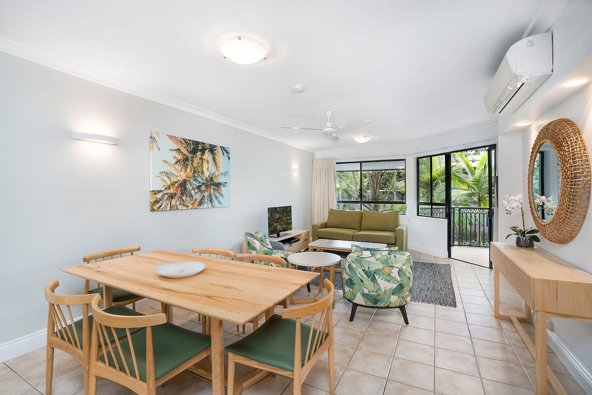 A modern living room with a wooden dining table, green cushioned chairs, a sofa, and a coffee table. There is a painting on the wall, a ceiling fan, and an air conditioning unit.