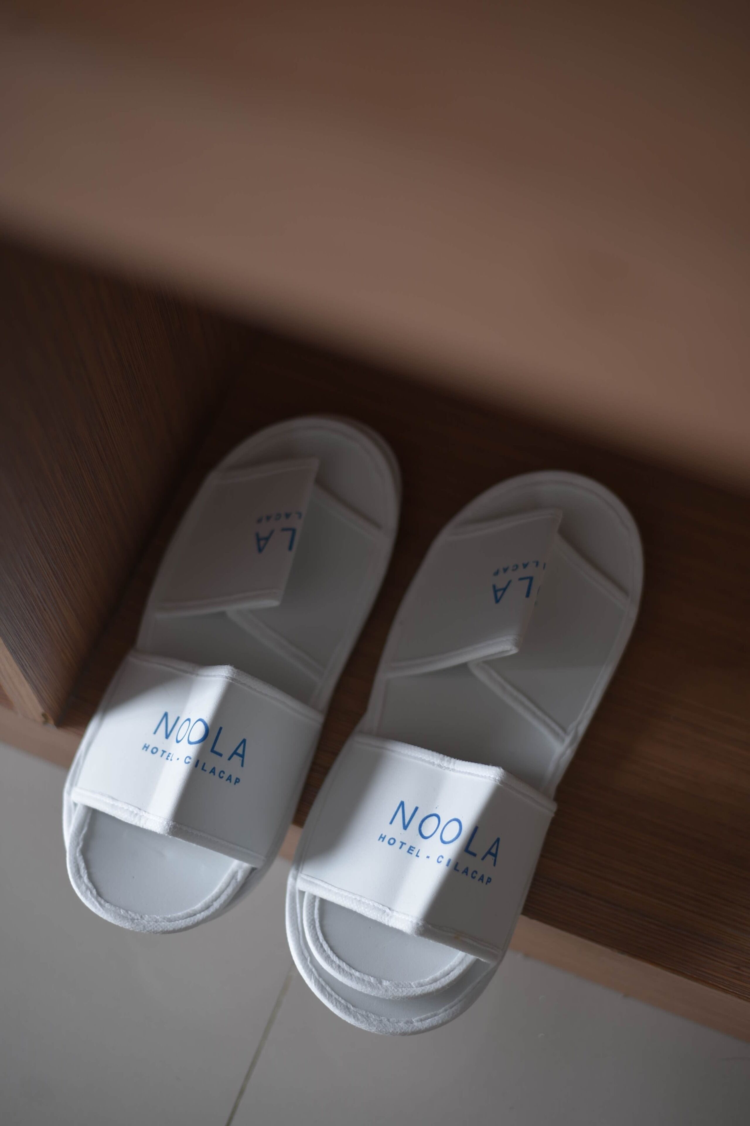 A pair of white Noolia slippers with the hotel name visible on the strap.